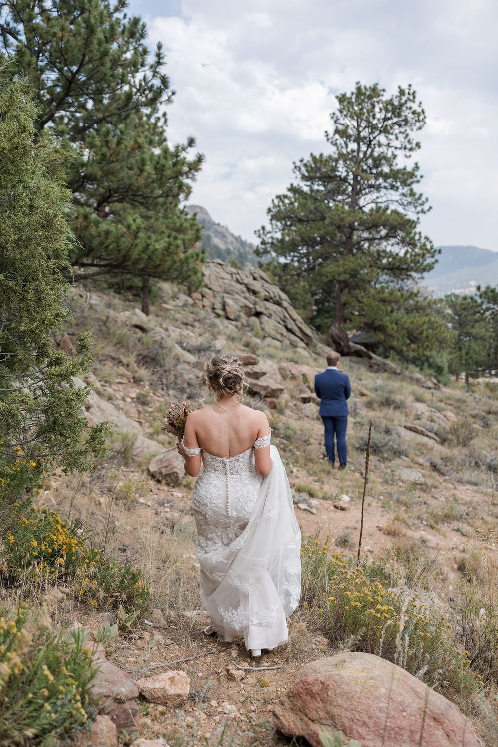 A wedding dress flows gracefully as someone walks along a rocky mountain trail surrounded by pine trees.