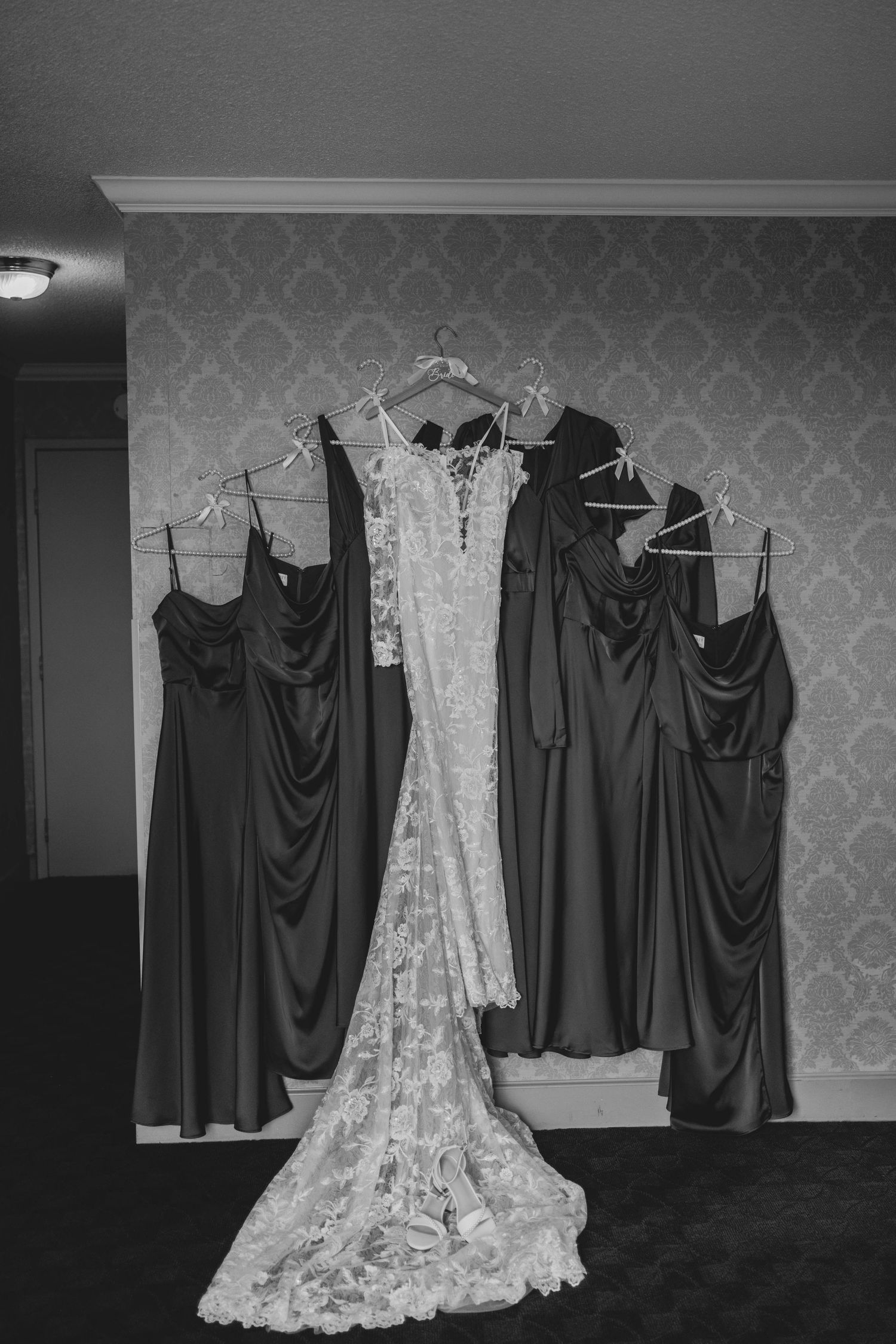 Wedding dresses hanging on display with a white lace gown and dark bridesmaid dresses against a patterned wallpaper.