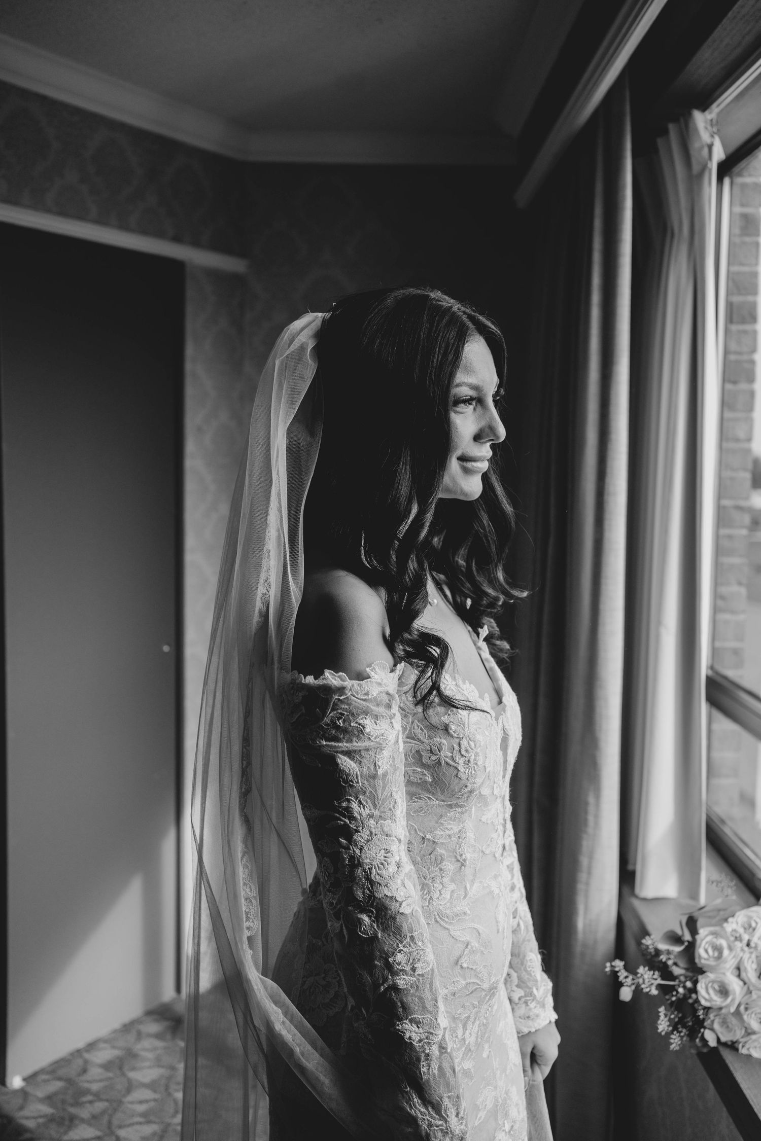 Artistic black and white series of a bride in lace wedding dress standing by windows with flowing veil.
