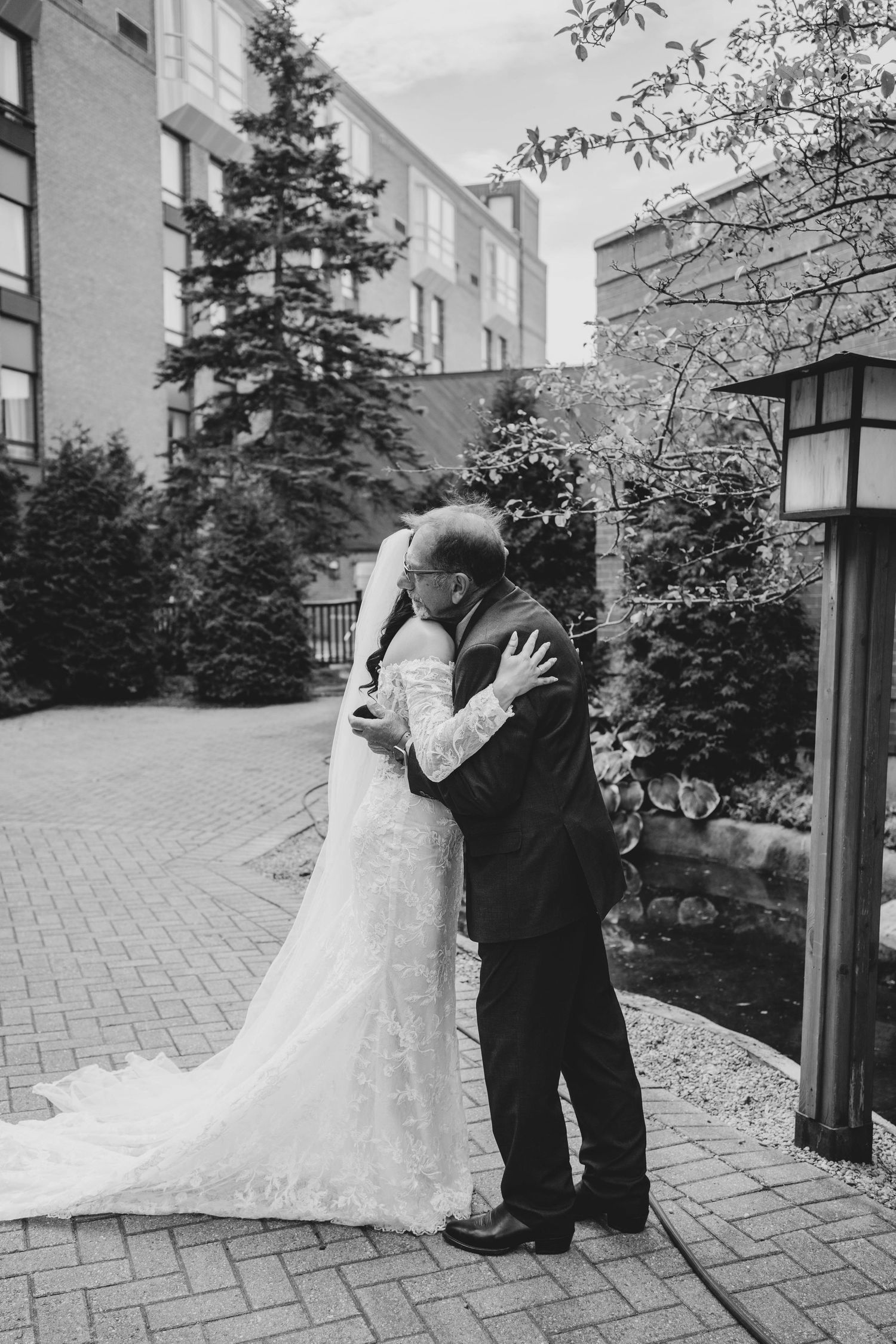 A romantic black and white wedding photo showing a couple embracing on a brick paved path surrounded by evergreen trees.