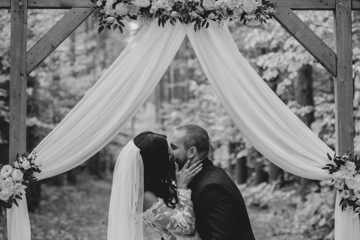 Elegant outdoor wedding ceremony setup with white fabric draping and floral decorations under a wooden pergola.