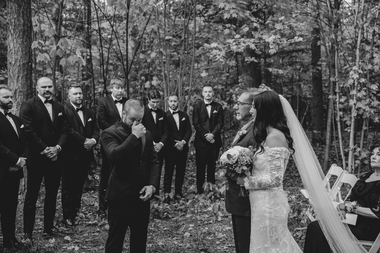 A group of wedding guests and the wedding party celebrate outdoors in a wooded area during a fall ceremony.
