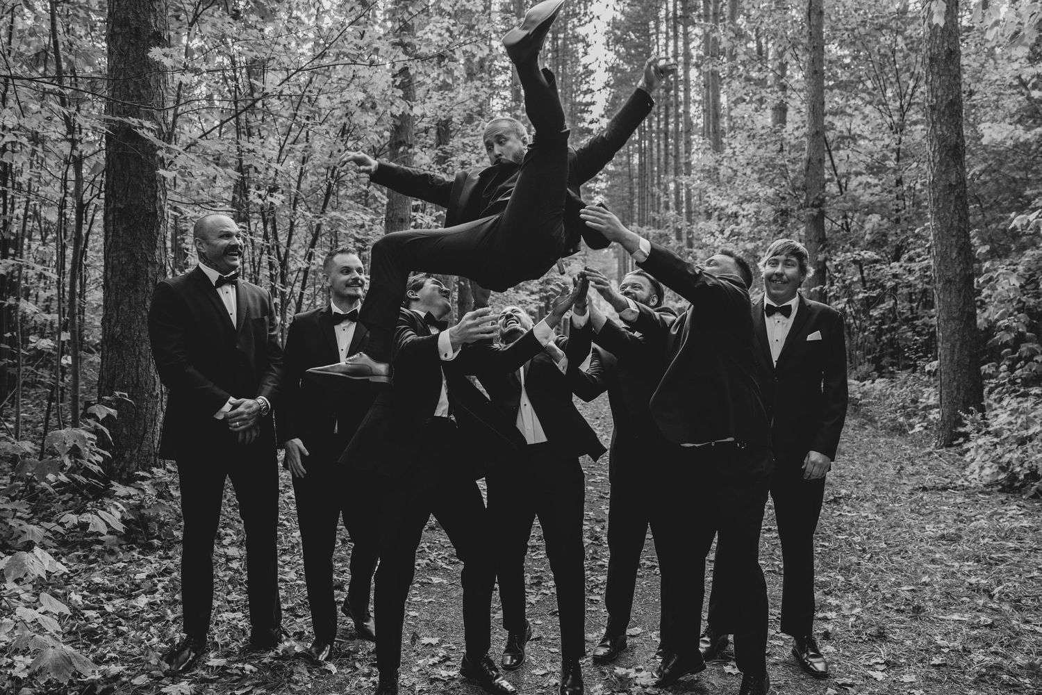 Wedding party in formal attire poses together in a wooded outdoor setting during autumn.