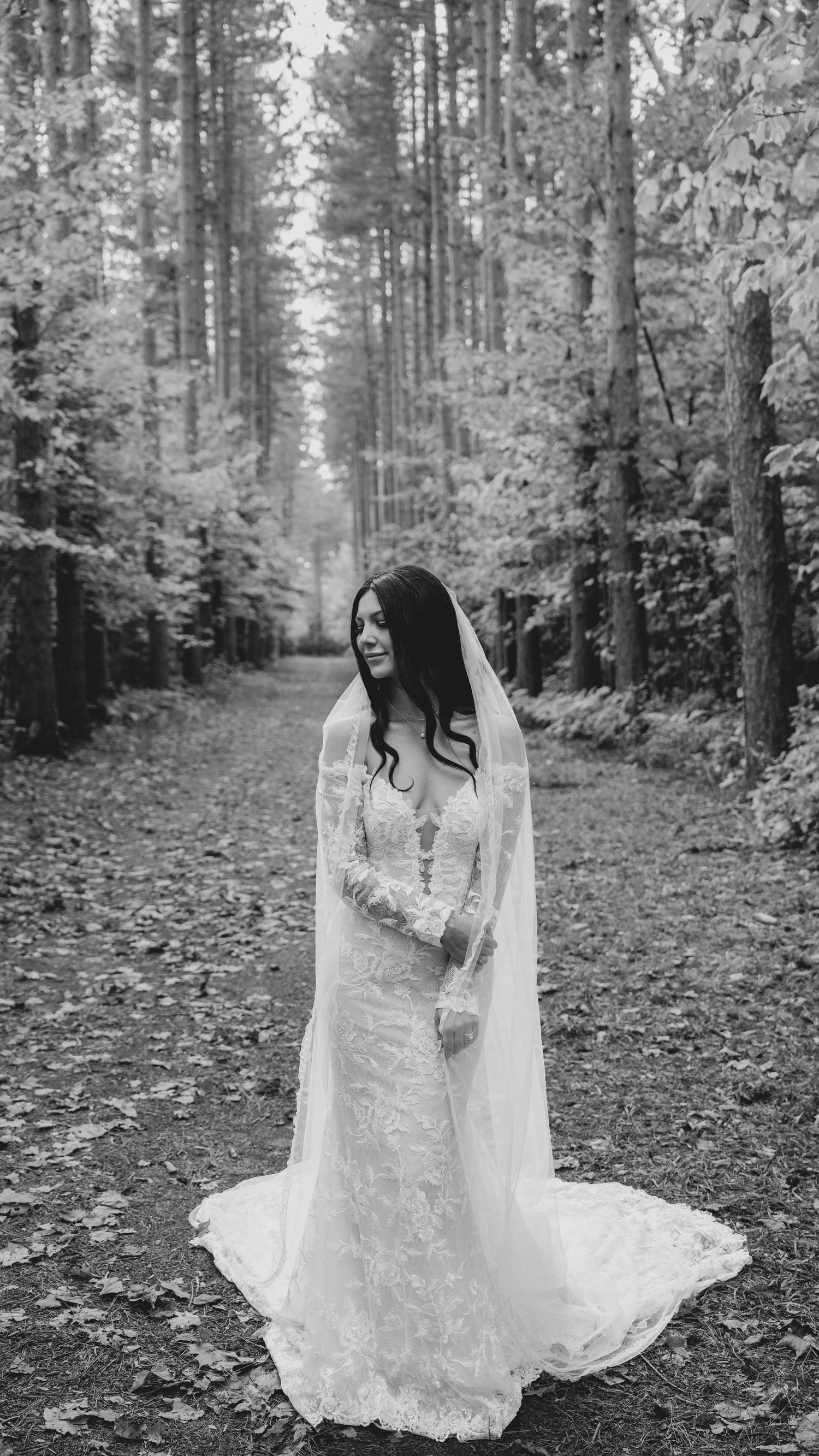 A bride in a long-sleeved lace wedding gown and veil stands in a serene forest path surrounded by tall trees.
