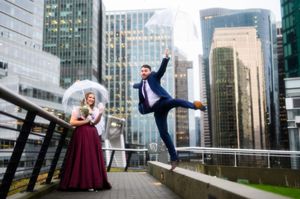 A couple in formal attire dances and poses together on a rooftop terrace with city skyscrapers in the background.