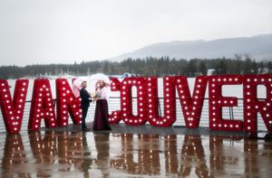 A large red sign with illuminated letters spelling out Vancouver on a waterfront with mountains in the background.