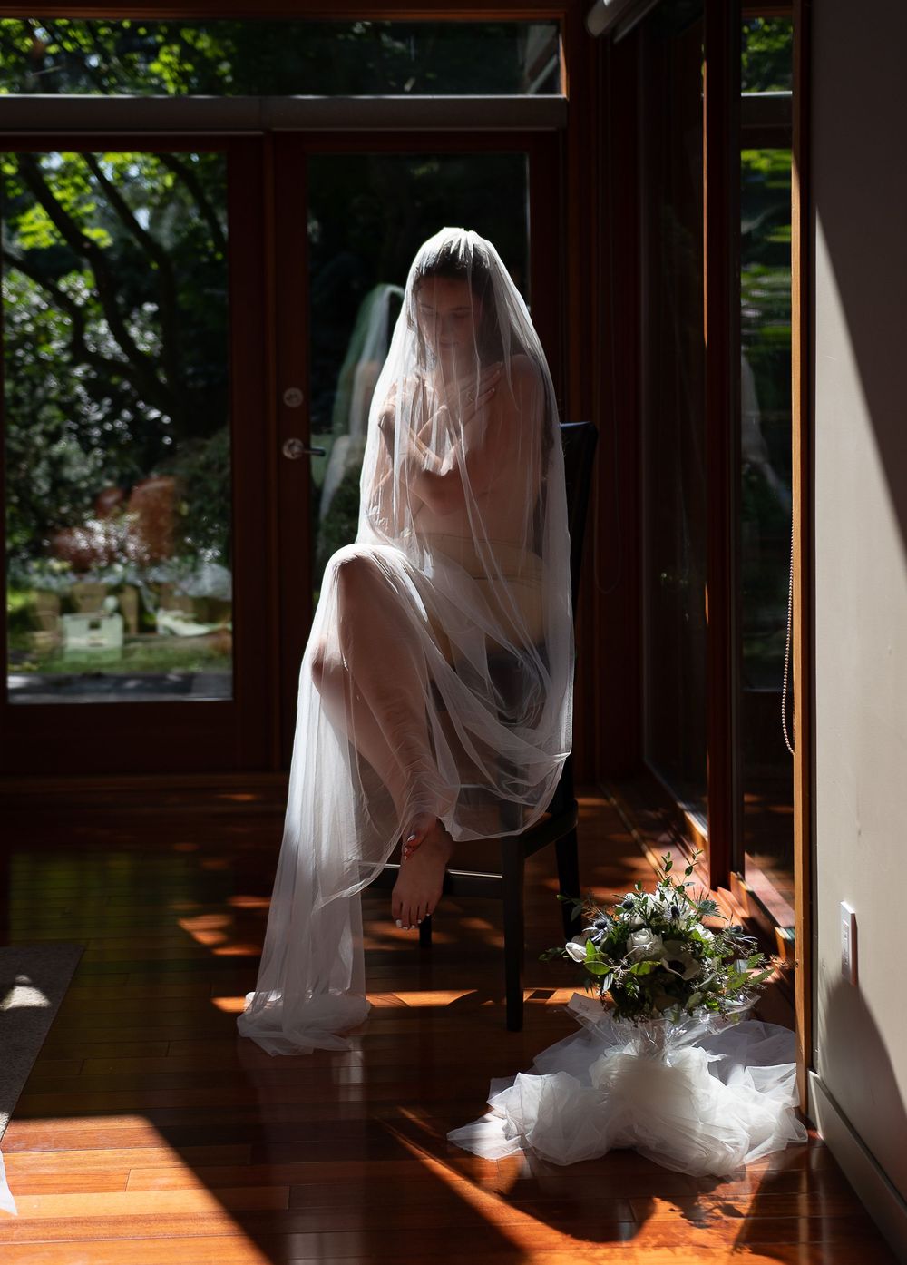 A bride in a flowing white dress and veil stands silhouetted against sunlit windows in a rustic wooden interior.