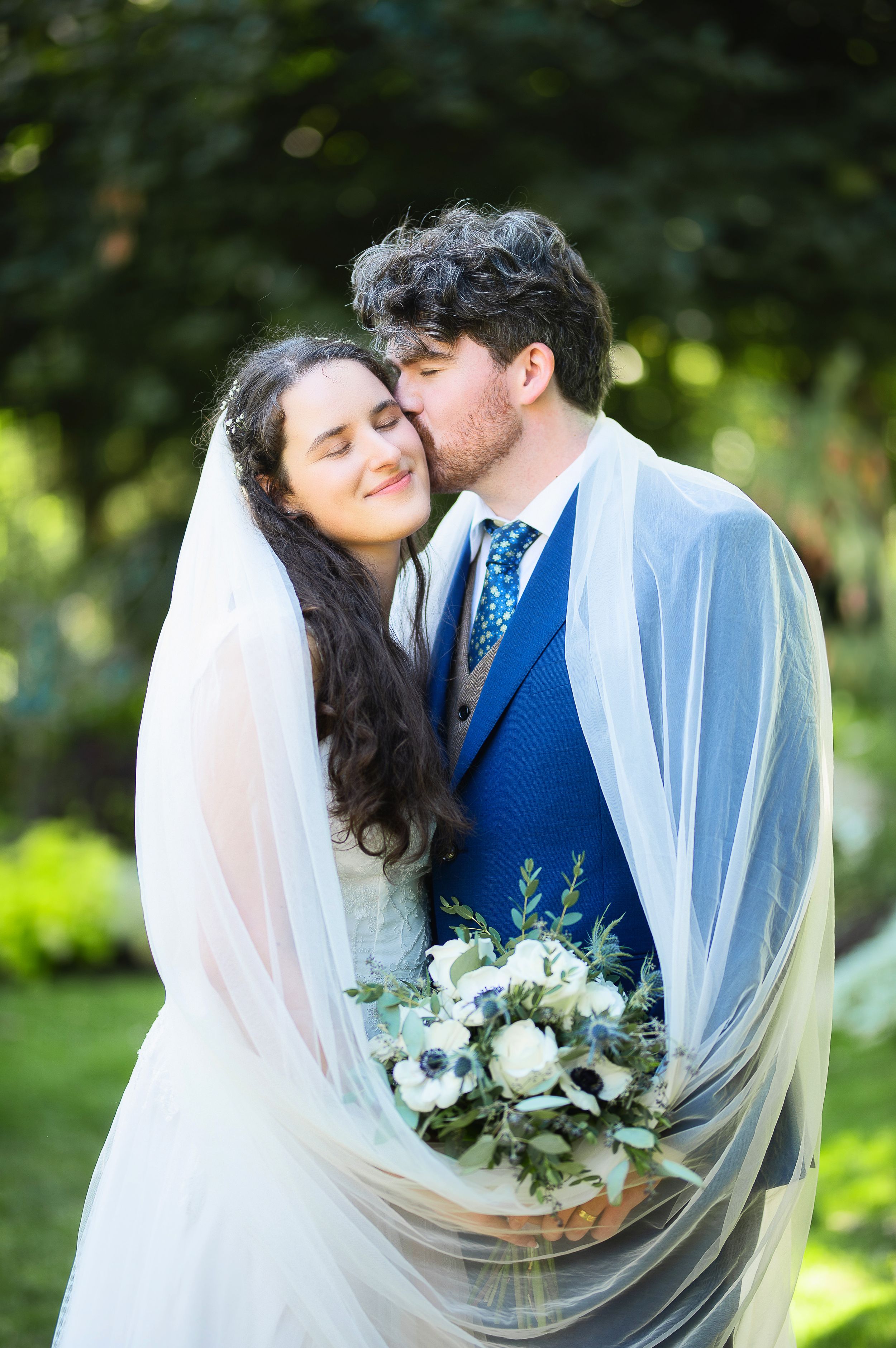 A romantic wedding couple embraces outdoors while holding a white and green floral bouquet.