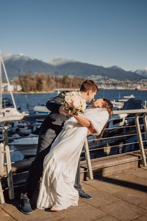 A couple in wedding attire performs a dip pose on a marina dock with mountains and boats in the background.