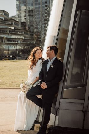 A couple in wedding attire share an intimate moment together outside a modern glass building.