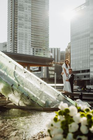 A couple poses elegantly by a modern glass building with white flowers in the foreground during their wedding photoshoot.