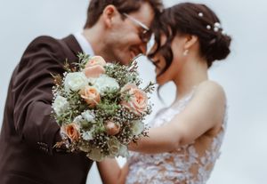 Romantic couple embraces during wedding ceremony with delicate peach and white floral bouquet details.