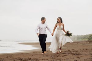 A couple in wedding attire walks hand in hand along a misty beach shoreline at sunset.