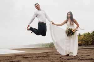 A couple in wedding attire jumps joyfully on a scenic beach with palm trees in the background.