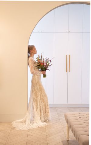 A figure in a flowing lace wedding gown holds a vibrant floral bouquet against a minimalist white interior.