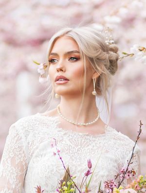 Ethereal bridal portrait with delicate lace dress and pearl necklace against soft pink cherry blossoms in spring.