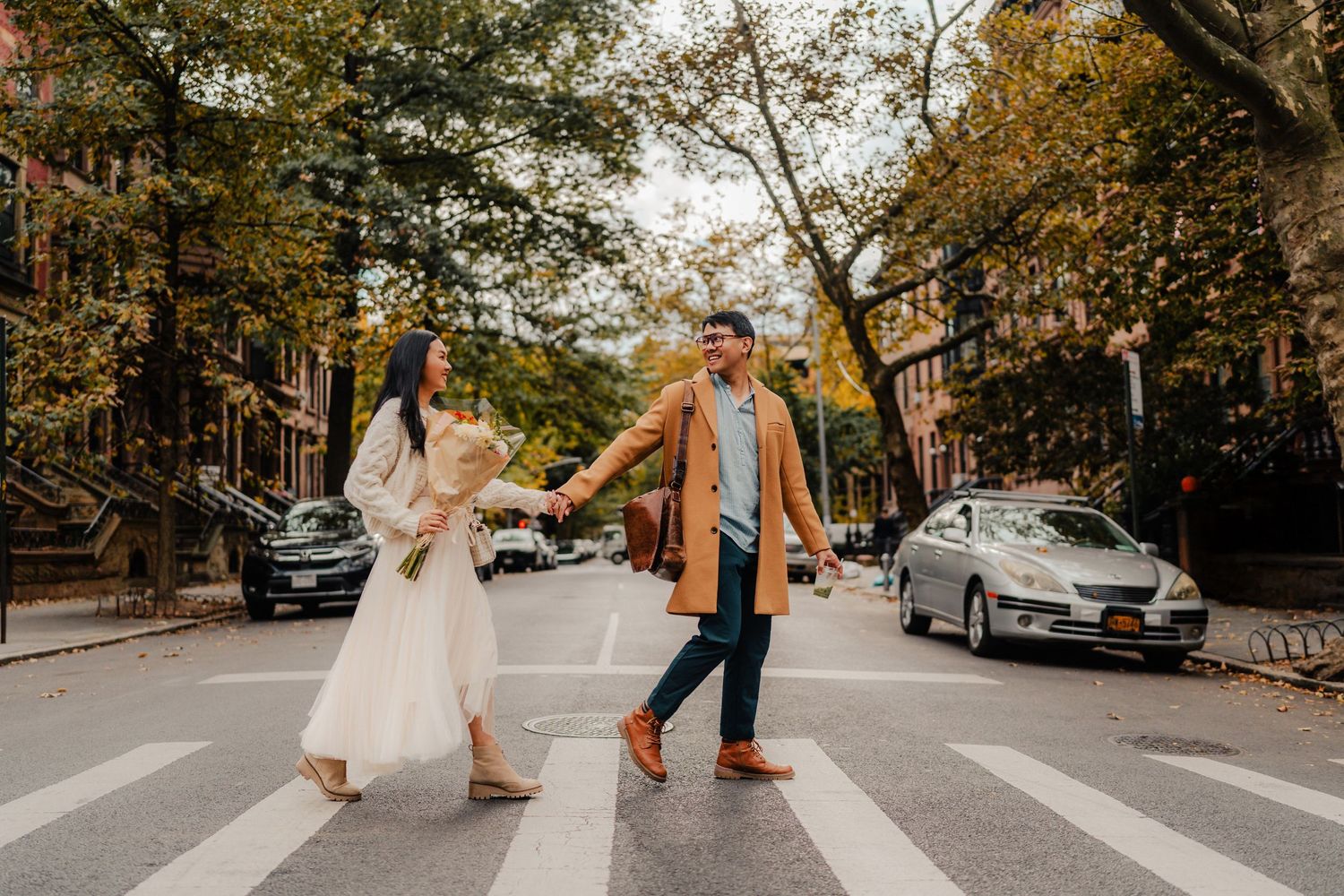 A couple holds hands while walking down a tree-lined autumn street with golden leaves falling around them.