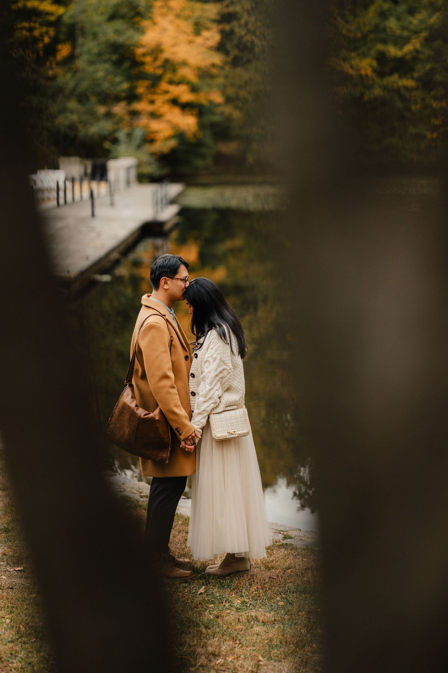 A couple in autumn attire shares an intimate moment among fall foliage and a serene lake backdrop.