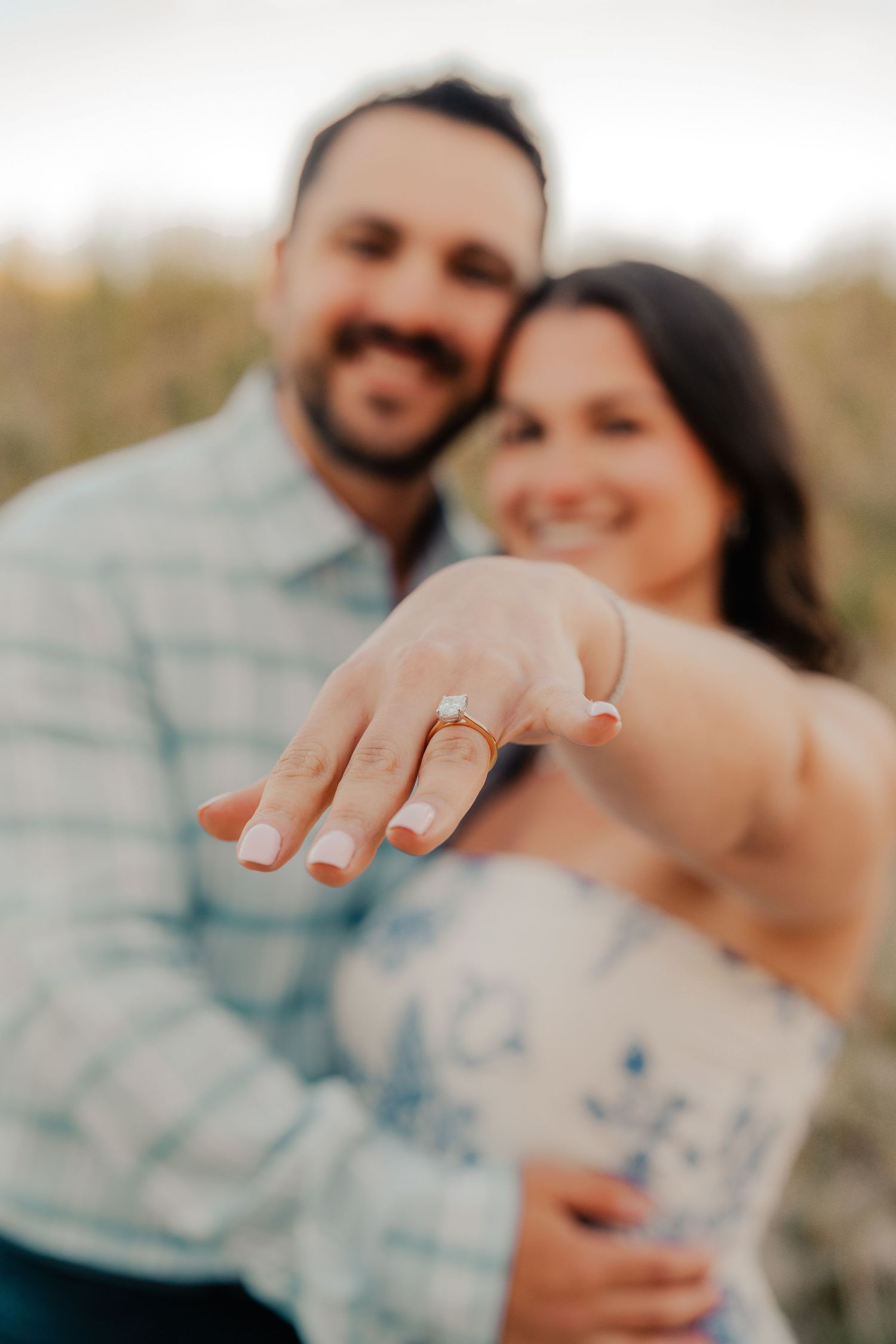 Smiling couple showing off engagement ring with diamond solitaire in outdoor setting.
