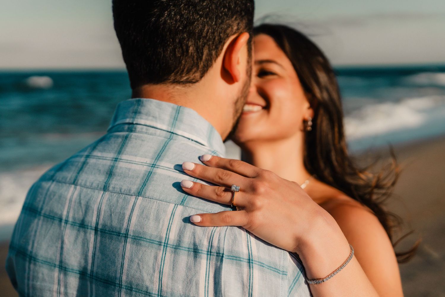 Couple sharing a romantic moment on the beach, woman smiling with engagement ring visible on her hand.