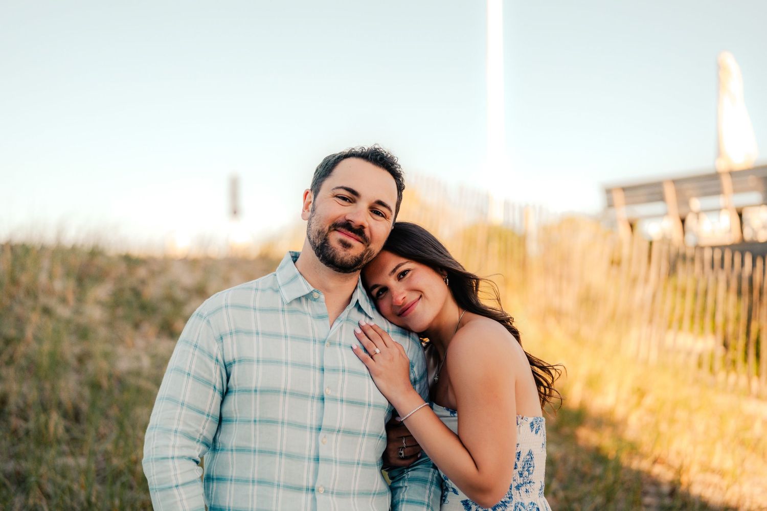Smiling couple posing outdoors at golden hour, woman resting head on mans shoulder near a wooden fence.
