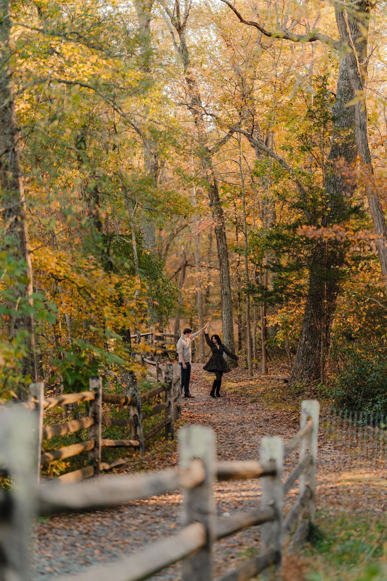 Two people walk along a leaf-covered forest trail surrounded by golden autumn trees and a rustic wooden fence.