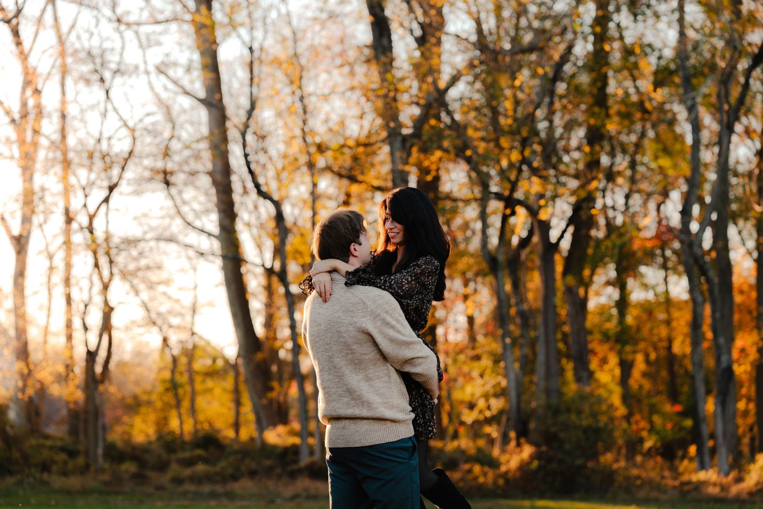 A couple embraces during a romantic autumn photoshoot in a wooded area with golden sunlight filtering through trees.