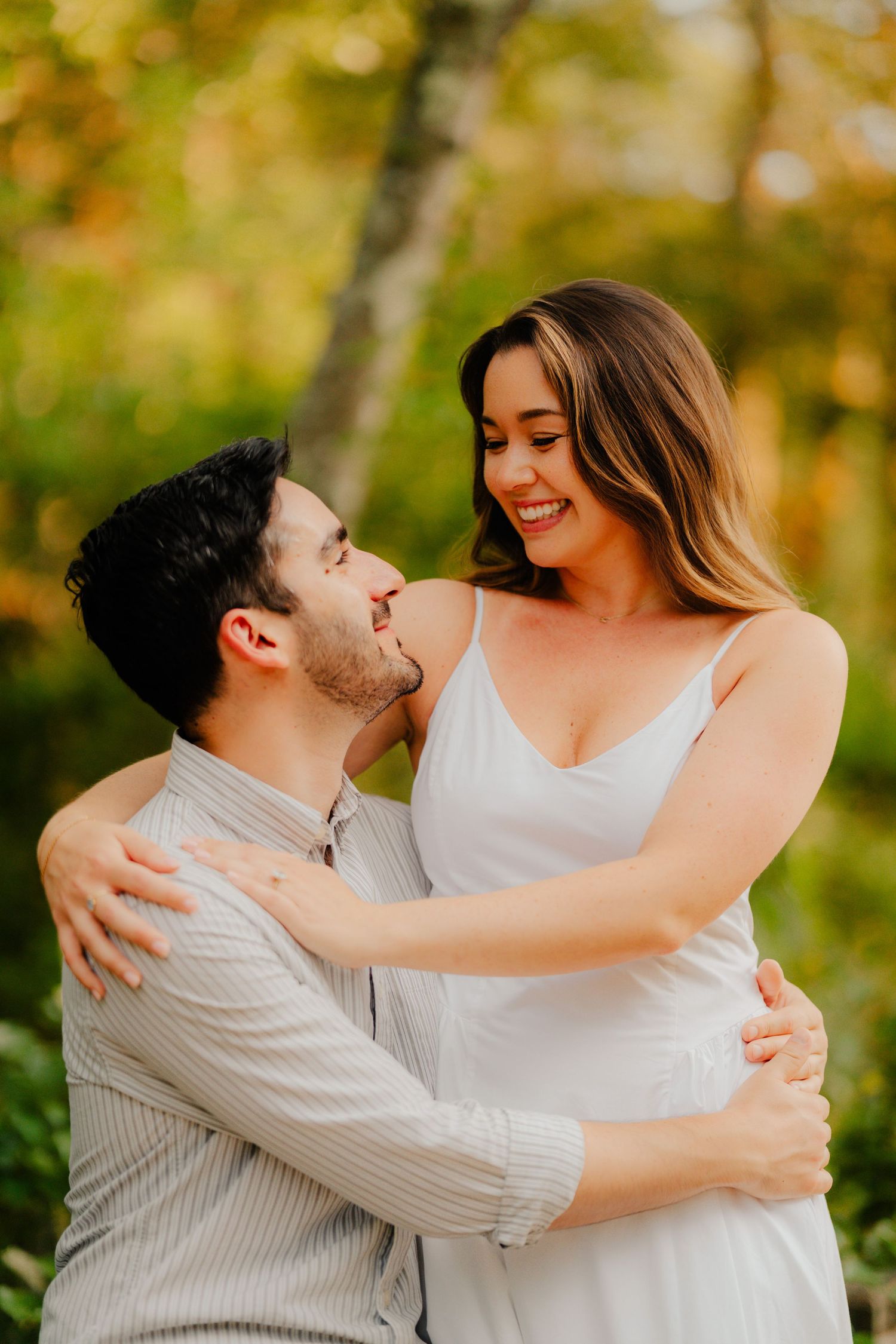 A romantic couple embraces and laughs together during an outdoor autumn photoshoot with golden foliage in the background.