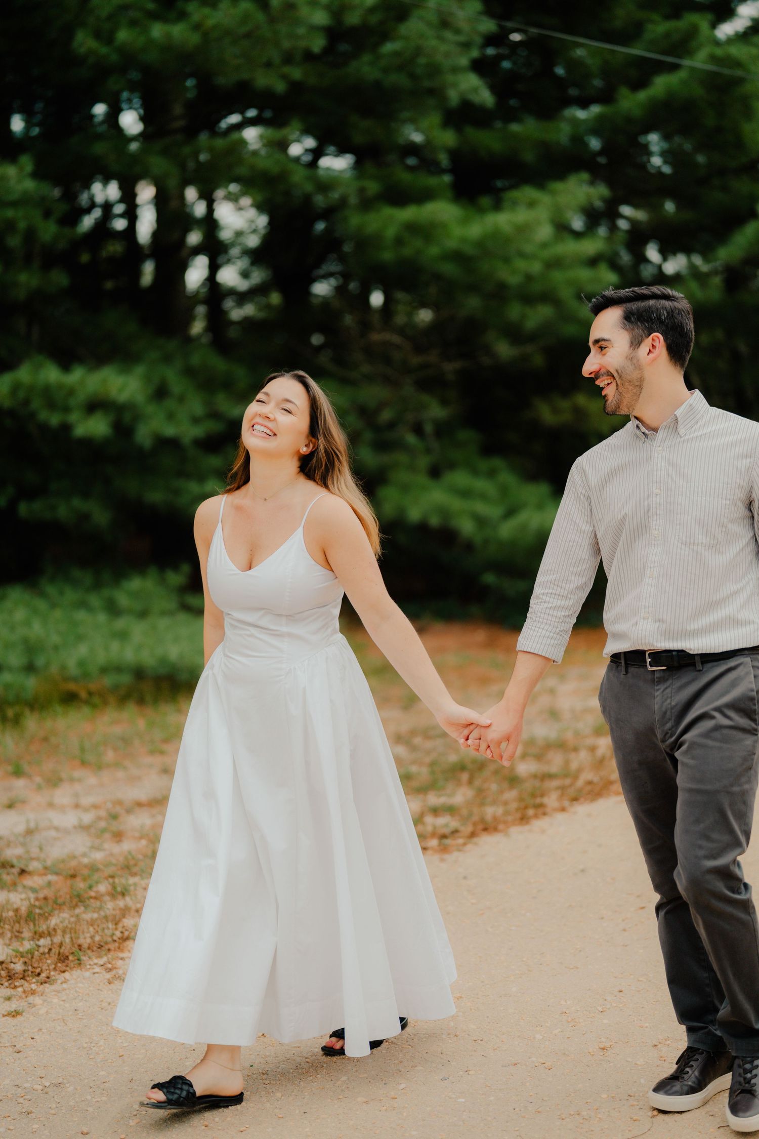A couple in wedding attire shares a joyful dance moment on a sandy path surrounded by green pine trees.