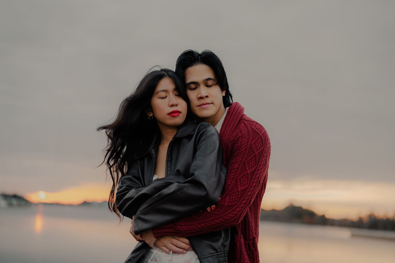 A young couple embraces tenderly at sunset by a lake, the woman in gray leather and man in a red knit sweater.