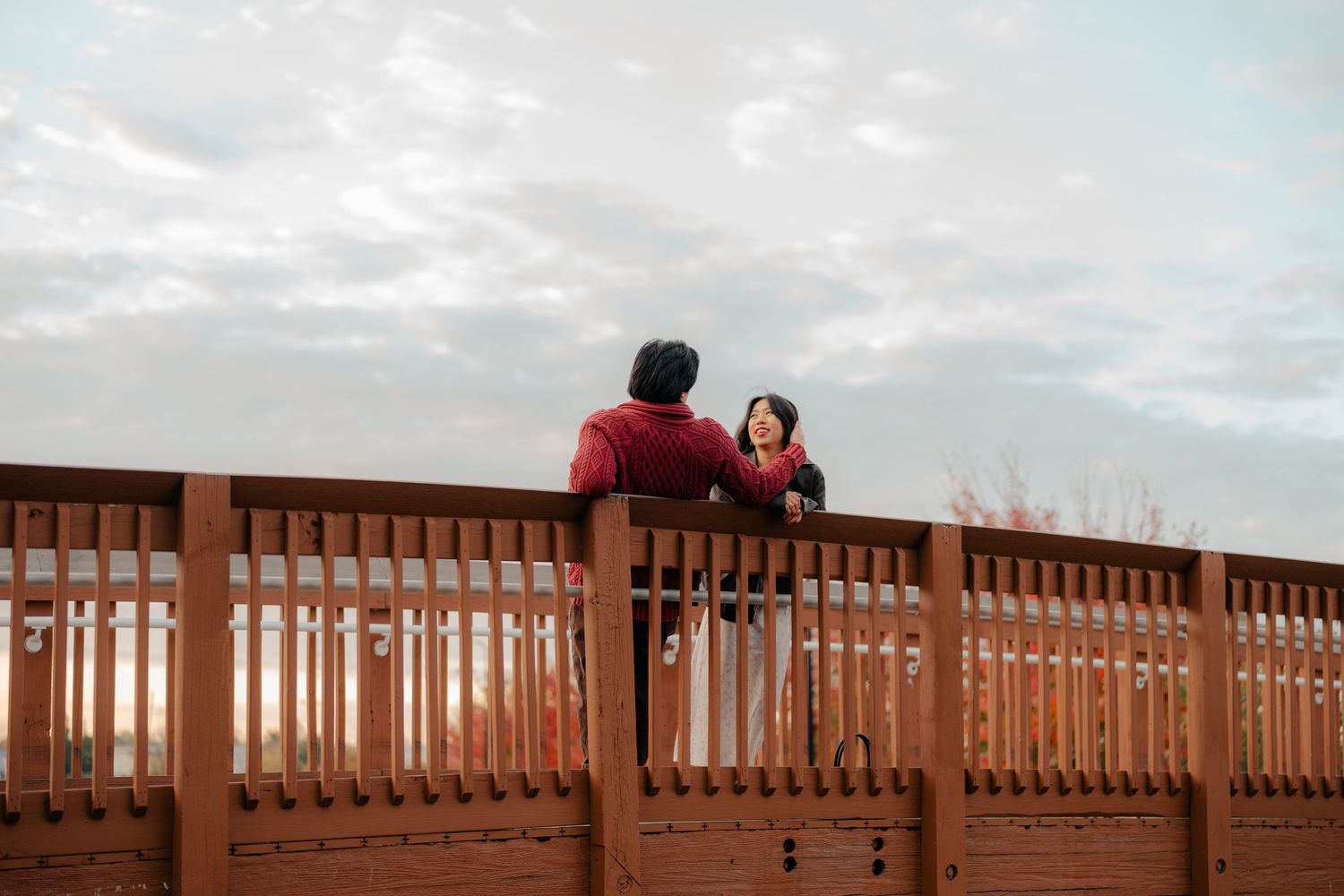 Two people leaning on a wooden bridge railing, sharing a conversation under a cloudy sky at dusk.