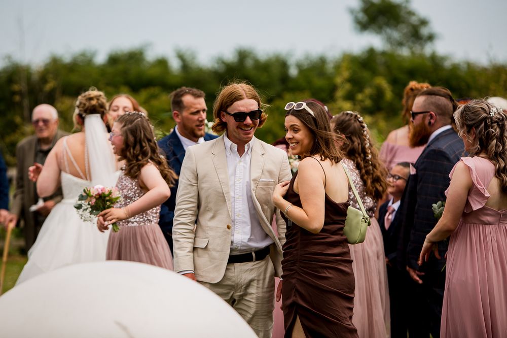 Wedding guests in formal attire gather outdoors on a sunny summer day.