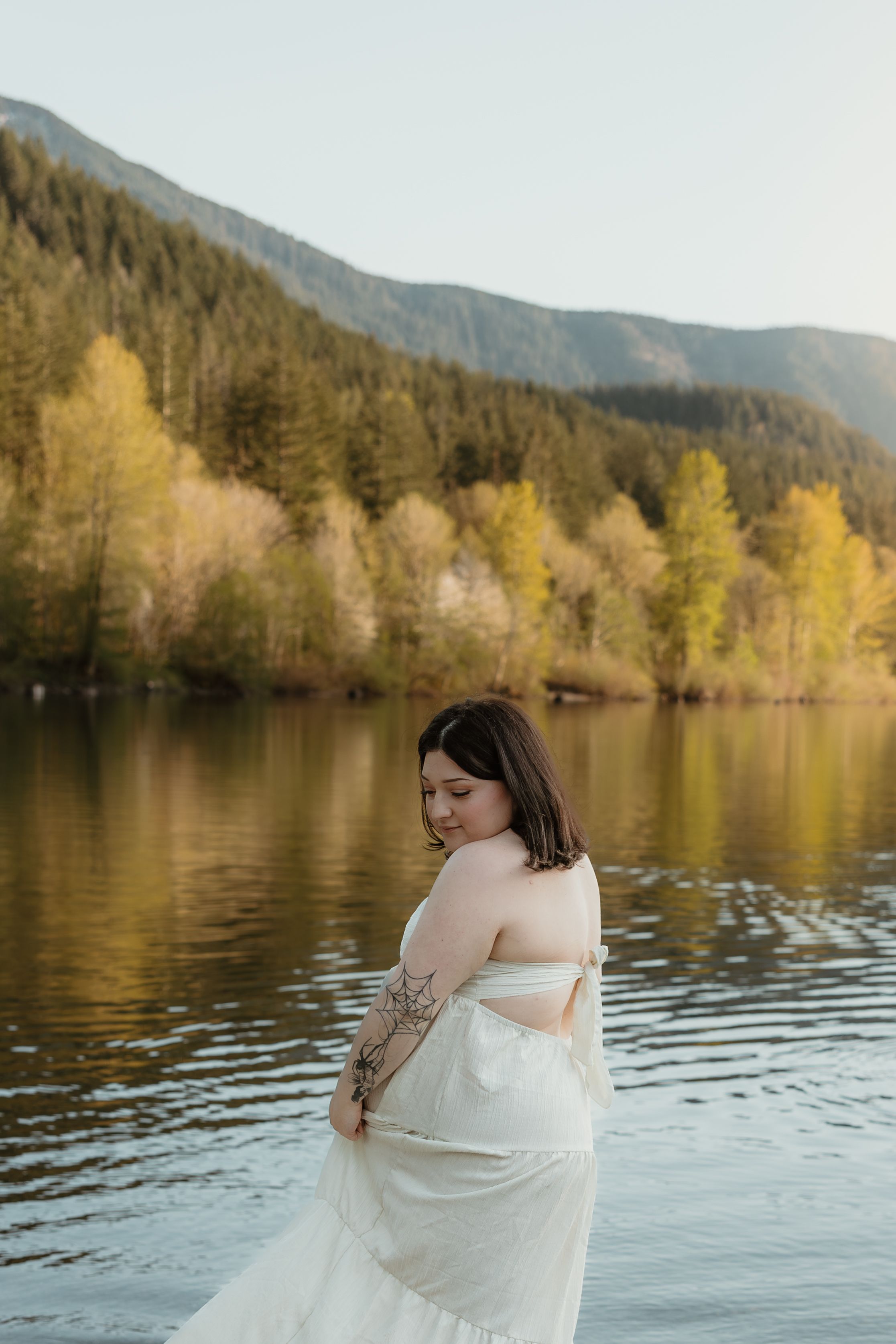 Woman in white dress stands by serene mountain lake with autumn trees reflecting in rippling water.