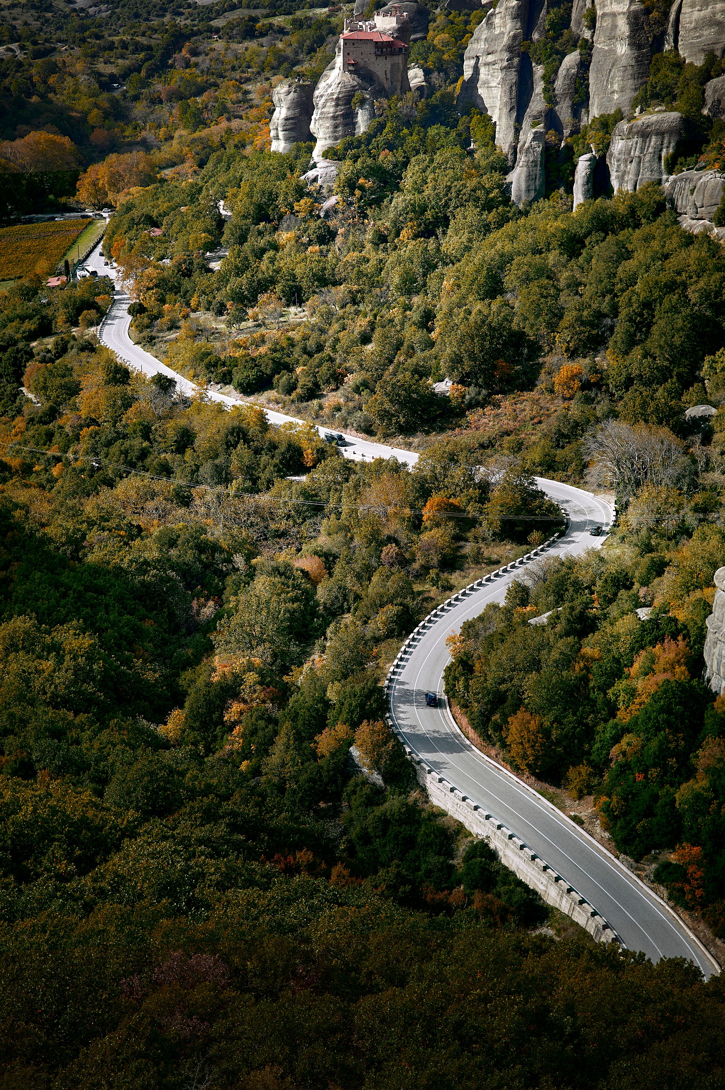 Aerial view of a winding mountain road cutting through dense autumn forest with cars traveling along its curves.