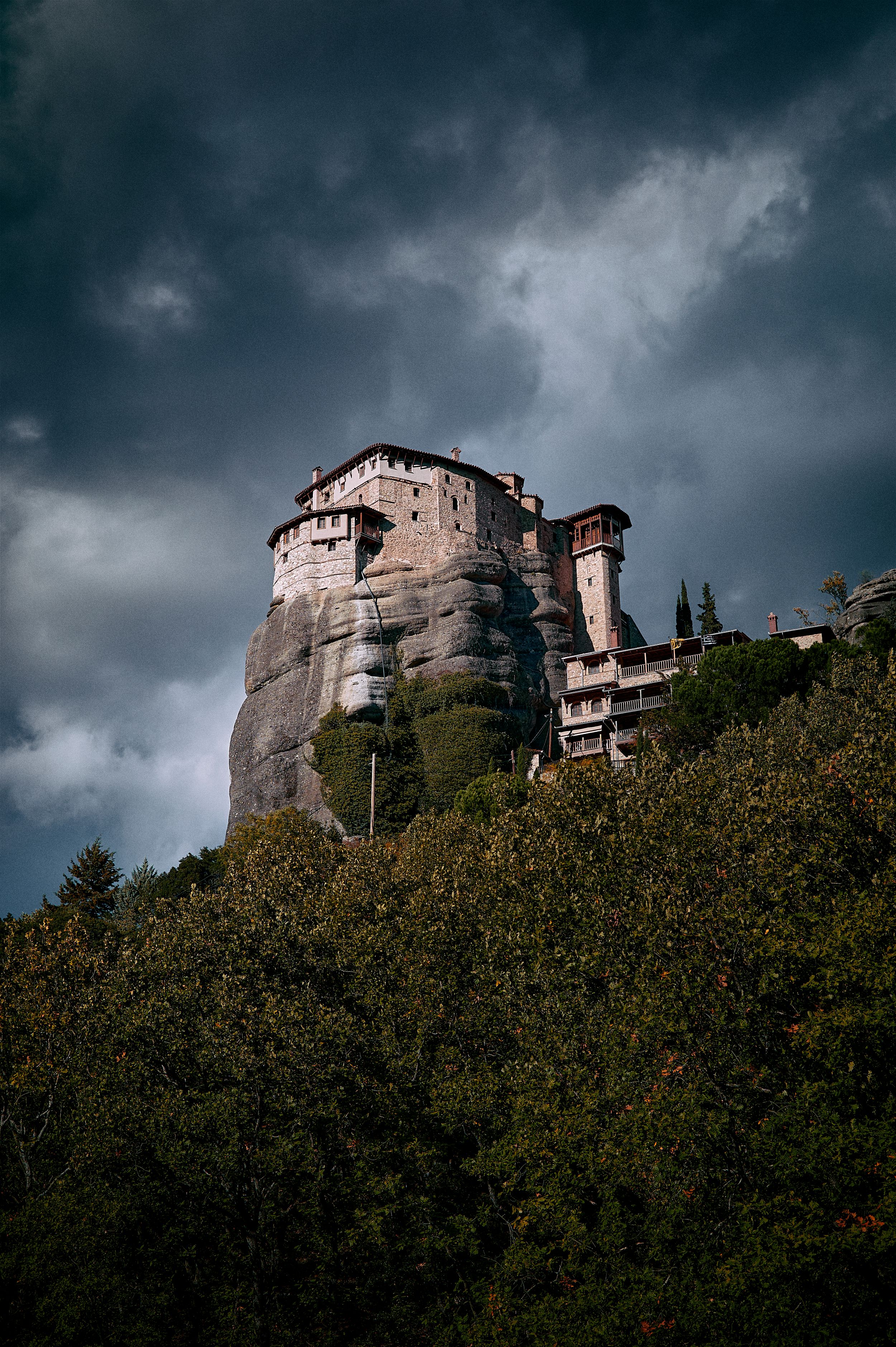 Ancient stone monastery perched on dramatic cliffs in Meteora, Greece under moody storm clouds.