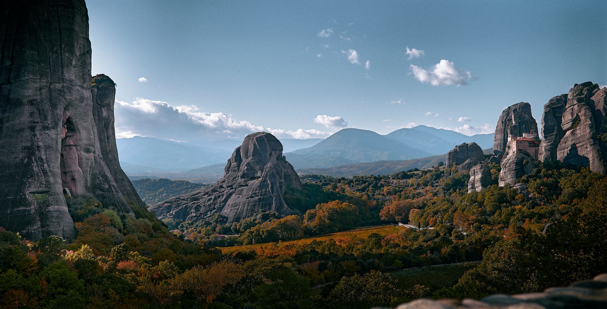 Panoramic mountain landscape with towering rock formations and autumn foliage against a blue sky with scattered clouds.