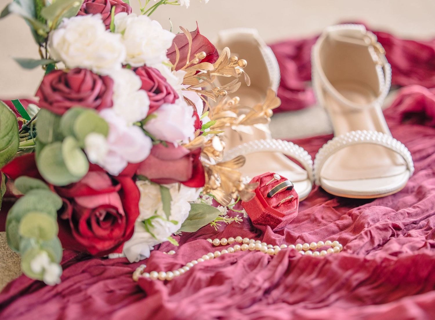 Wedding details with roses and pearls arranged on burgundy fabric.