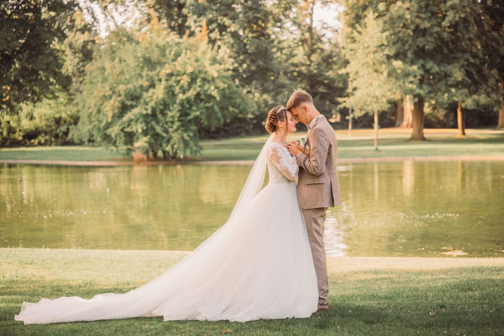 A romantic wedding couple embraces by a tranquil lake in a scenic park setting during golden hour.