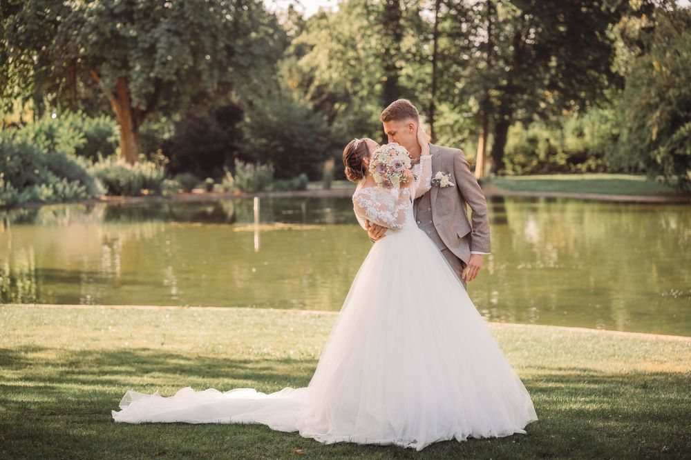 A romantic wedding couple embraces by a serene pond in a lush garden setting during golden hour.