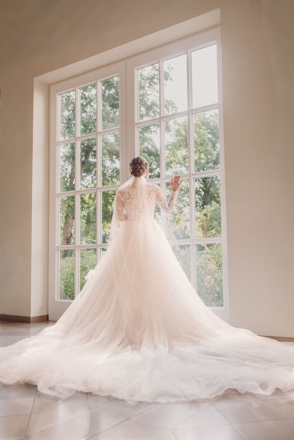 A flowing white wedding gown with lace details and long train photographed from behind against a large bright window.
