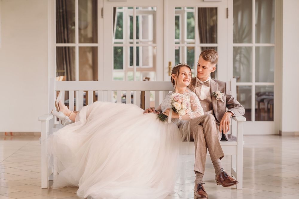 A happy couple in wedding attire sits together on a white floor in an elegant bright room with large windows.