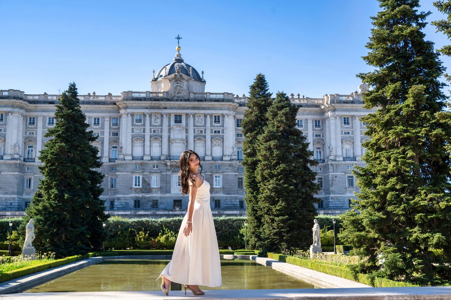 A grand palace facade with white architecture and evergreen trees lines a formal courtyard walkway.