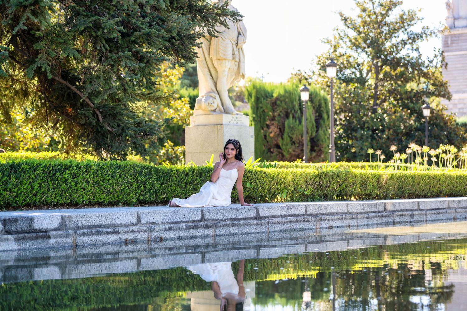 A person in white dress sits by a reflective pond near manicured hedges and garden statuary on a sunny day.