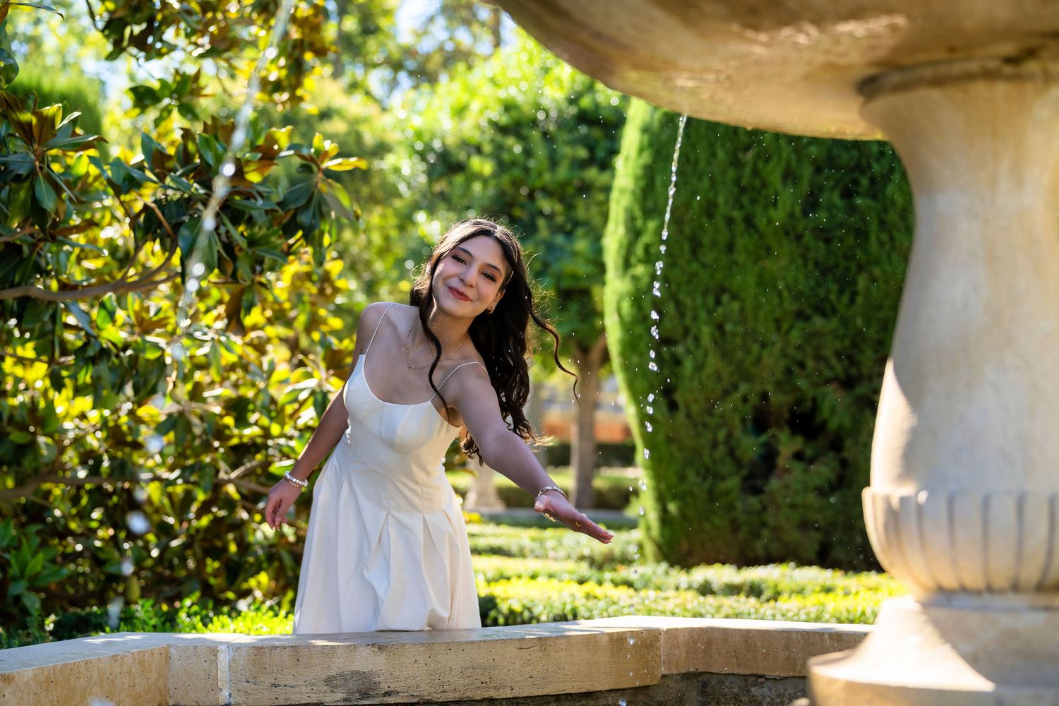 A person in a white dress poses gracefully in a sunlit garden setting with ornate architectural elements.