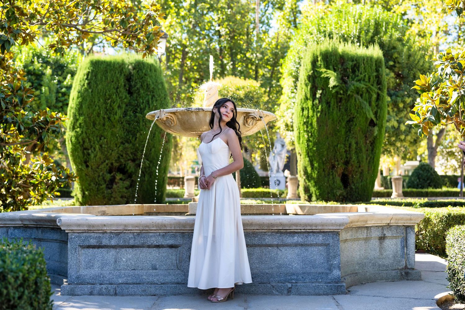 A person in a flowing white dress and sun hat stands by an ornate stone fountain in a manicured garden with topiary.