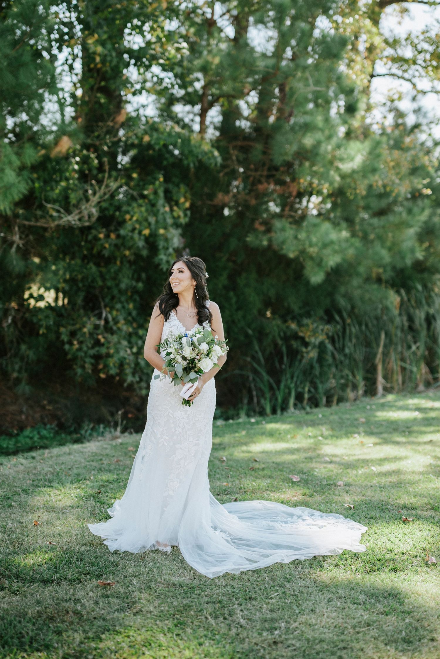A bride in a flowing white wedding dress poses gracefully on a sunlit grassy lawn surrounded by trees and greenery.