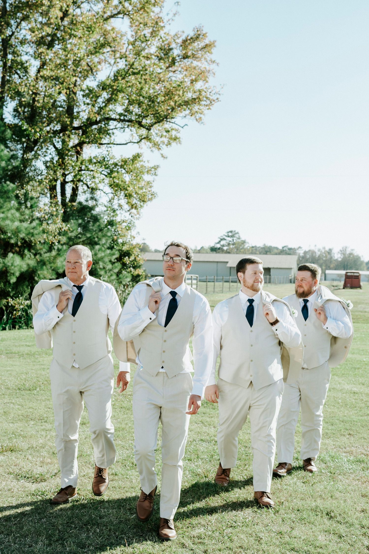 Groomsmen in white suits run playfully across a sunny green lawn during a wedding photoshoot.
