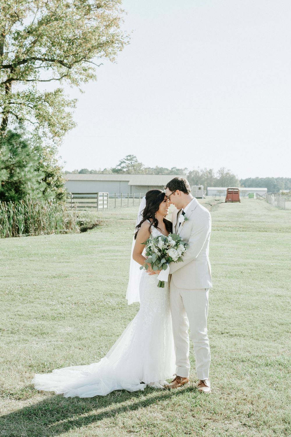 A romantic wedding couple shares an intimate moment in a sunlit field with trees and fences in the distance.