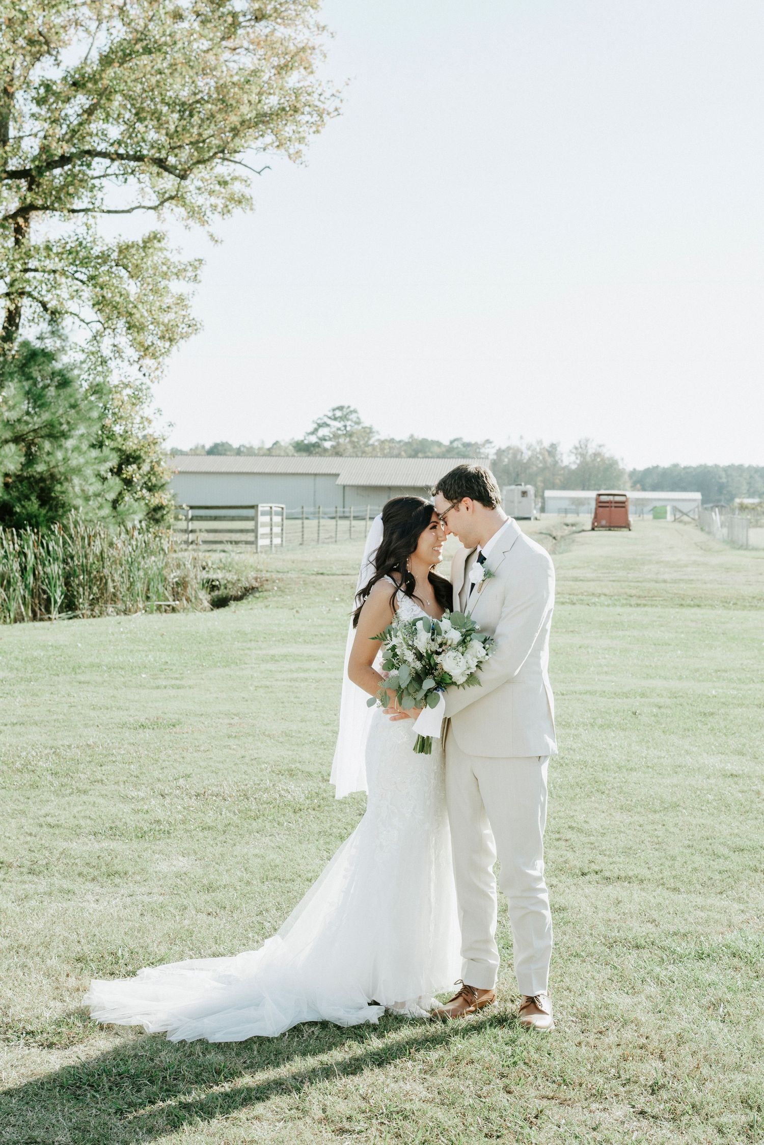 A romantic wedding couple shares an intimate moment in a sunlit field with trees and fences in the distance.