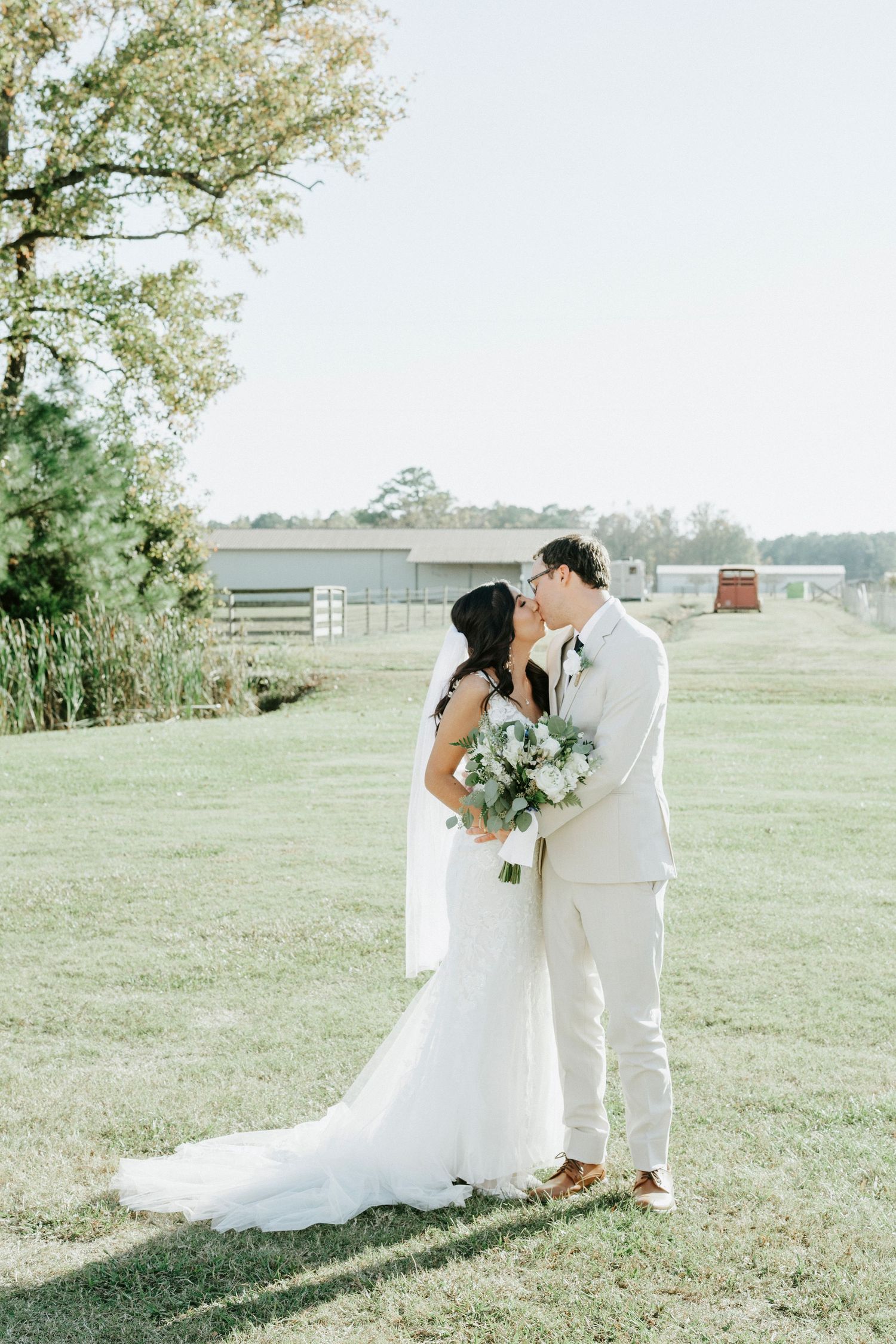 A romantic wedding couple shares intimate moments in a sunlit countryside setting with white attire and floral bouquet.